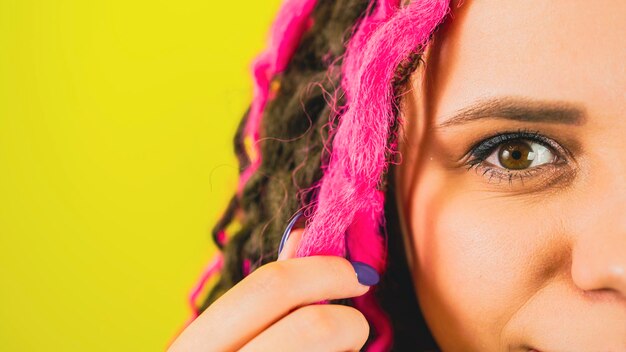 Молодая женщина скручивает розовые волосы и смотрит в камеру на желтом фоне. Часть женского лица с вьющимися дредами, выражающими положительные эмоции.