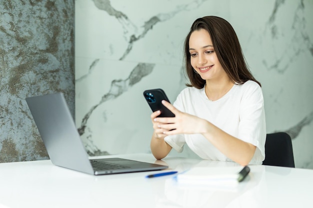 Young woman use a laptop to work in the kitchen writes something in a notebook with a pen and speaks on the phone