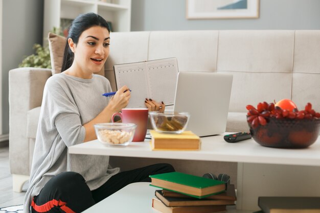 若い女性は、リビングルームのコーヒーテーブルの後ろの床に座っているノートにラップトップの保持とペンでポイントを使用しました