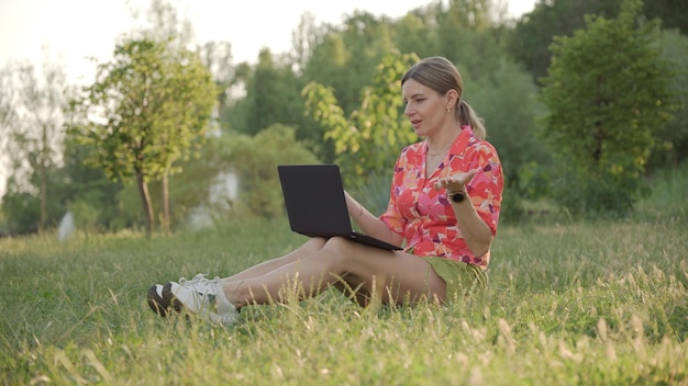 Young woman uses laptop sitting in park on grass