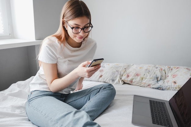 Young woman uses smartphone and laptop at home on the bed