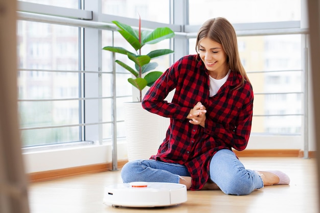 Young woman using automatic vacuum cleaner to clean the floor, controlling machine housework robot.