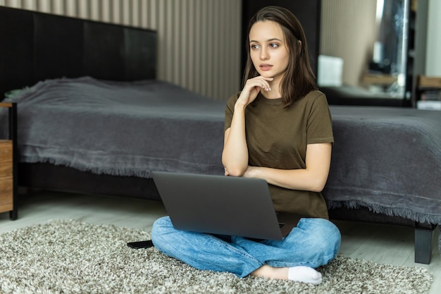 질문에 대해 생각하는 턱에 손으로 바닥에 앉아 컴퓨터 노트북을 사용하는 젊은 여자, 잠겨있는 표정. 사려깊은 얼굴로 웃고 있습니다.
