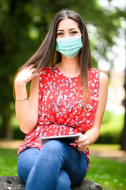 Young woman using a digital tablet sitting on a banch in a park during coronavirus times