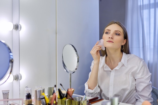 Young woman using gua sha massage scraper sitting in front of makeup mirror at home