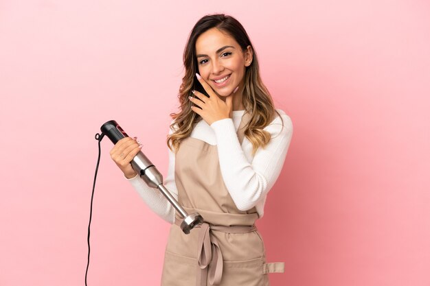 Young woman using hand blender over isolated pink background happy and smiling