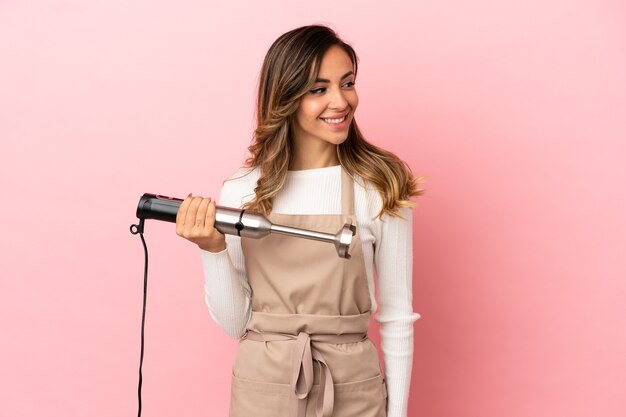 Young woman using hand blender over isolated pink background looking side