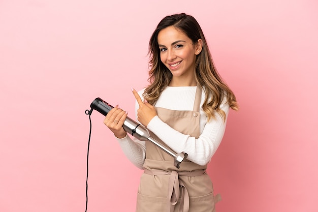 Young woman using hand blender over isolated pink background pointing to the side to present a product