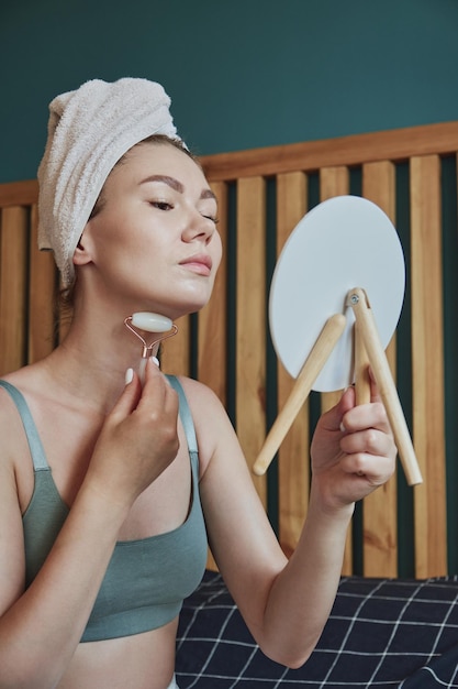 Young woman using jade facial roller for face and neck massage sitting on bed in bedroom looking in the mirror