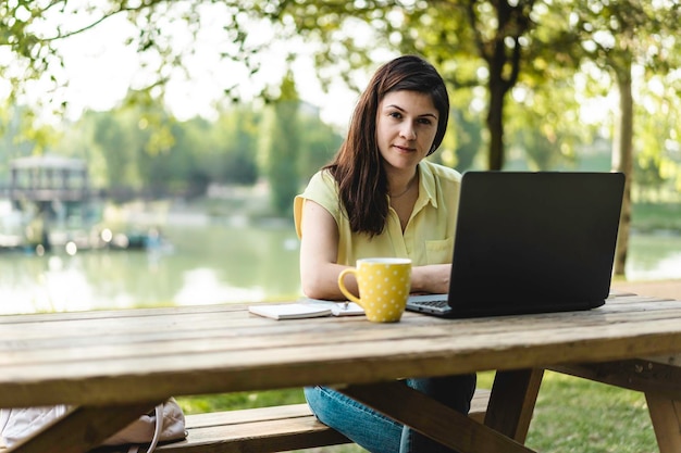 Giovane donna che utilizza un computer portatile nel parco pubblico millenaria freelance femminile che lavora in remoto e prende appunti in un parco cittadino concetto di tecnologia e lavoro a distanza utilizzo di dispositivi tecnologici all'aperto