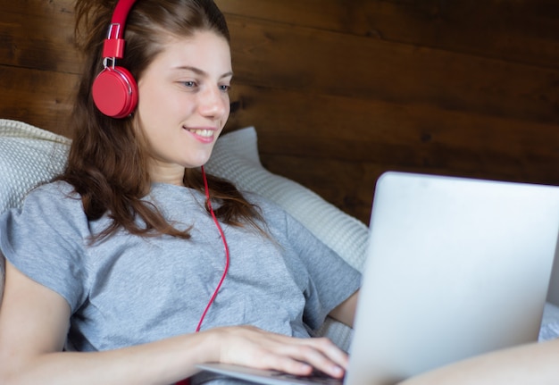 Young woman using laptop and headphones in bed.