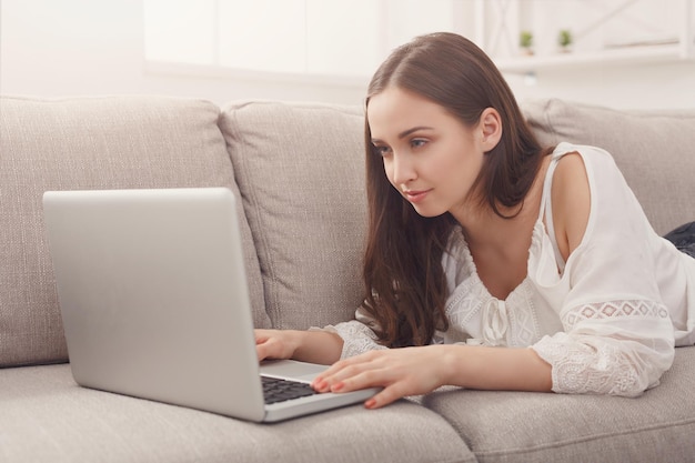 Young woman using laptop at home while lying on beige couch. Dark-haired girl in casual, copy space