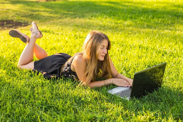 Young woman using laptop in the park lying on the green grass. Leisure time activity concept.