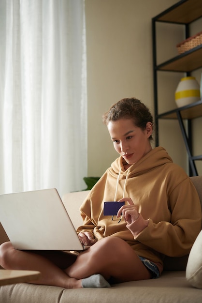 Young woman using laptop to pay online with her credit card while sitting on sofa in the room