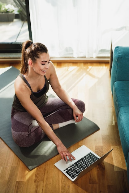 Young woman using laptop while doing stretching exercises at home in the morning.