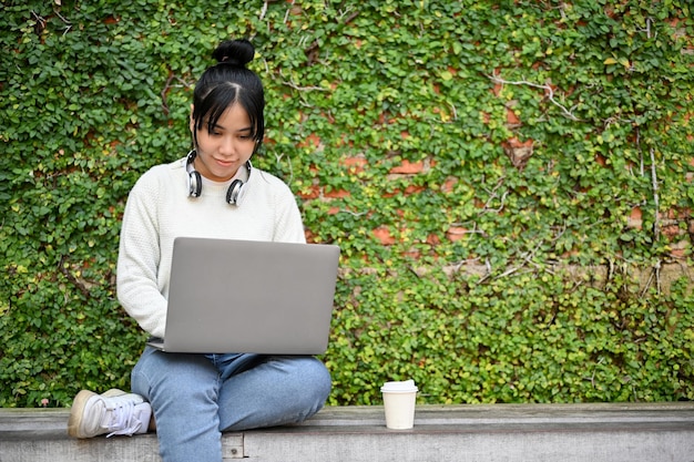 Young woman using laptop while sitting in the park with a cup of coffee beside her