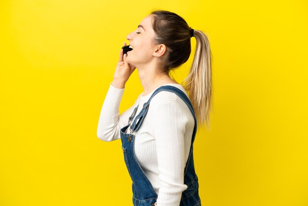 Young woman using mobile phone over isolated yellow background laughing in lateral position