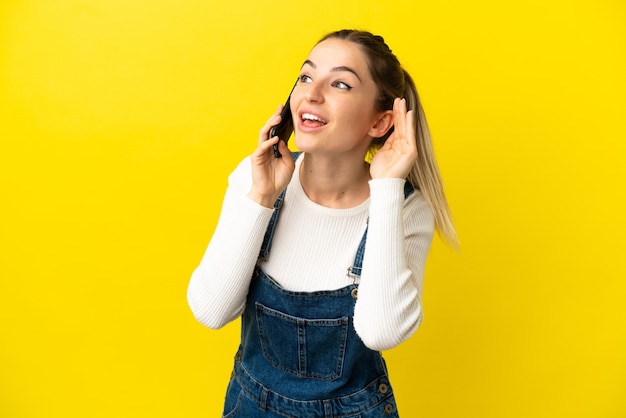 Young woman using mobile phone over isolated yellow background listening to something by putting hand on the ear