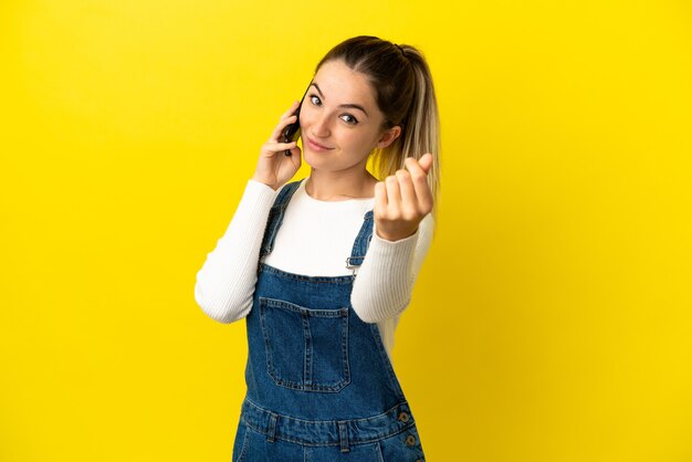 Young woman using mobile phone over isolated yellow background making money gesture