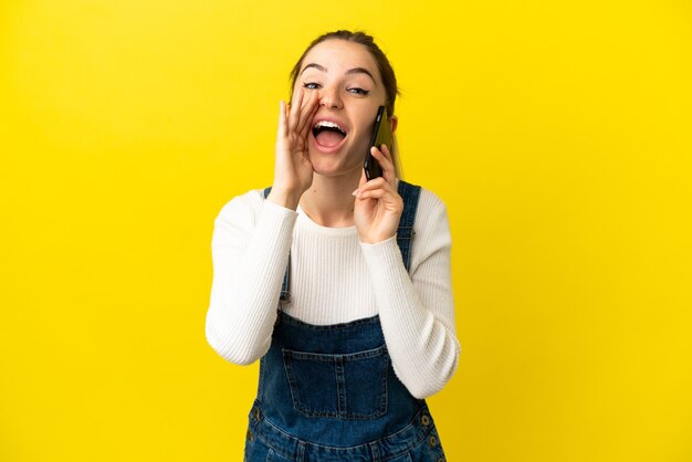 Young woman using mobile phone over isolated yellow background shouting with mouth wide open