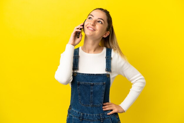 Young woman using mobile phone over isolated yellow background thinking an idea while looking up