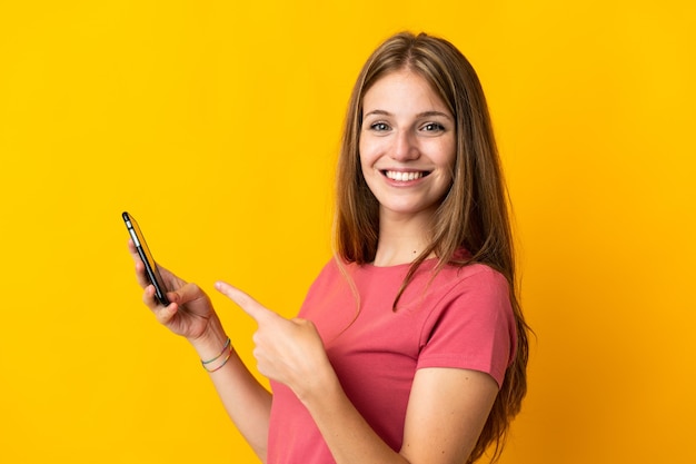 Young woman using mobile phone isolated on yellow and pointing it