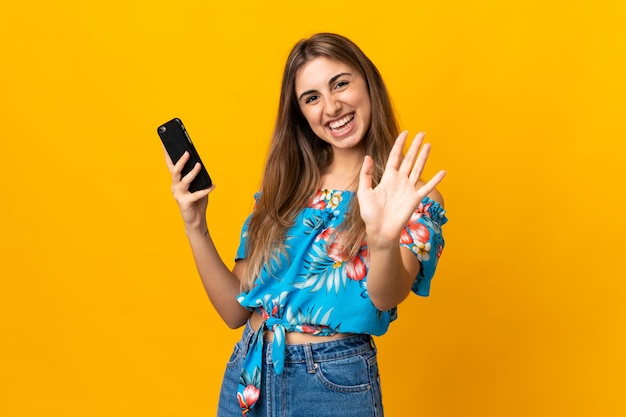 Young woman using mobile phone over isolated yellow saluting with hand with happy expression