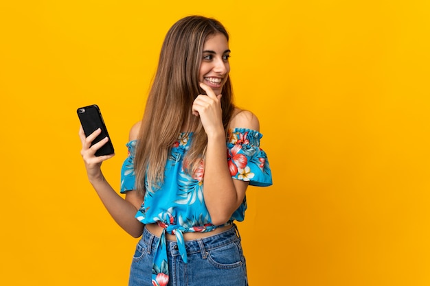 Foto giovane donna che per mezzo del telefono cellulare sopra giallo isolato che pensa un'idea e che guarda lato