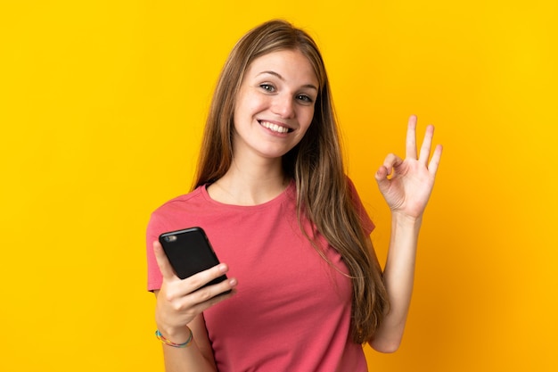 Young woman using mobile phone isolated on yellow wall showing ok sign with fingers