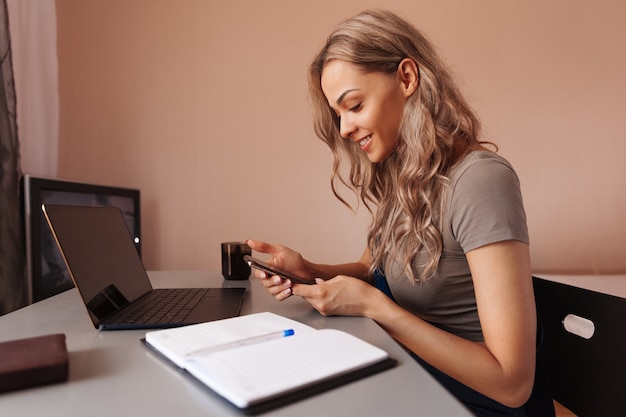 Young woman using mobile phone and smiling while sitting at table at home