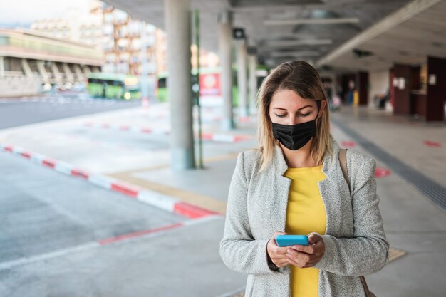 コロナウイルスの発生時にバス停で待っている間に携帯電話を使用している若い女性-顔に焦点を当てる