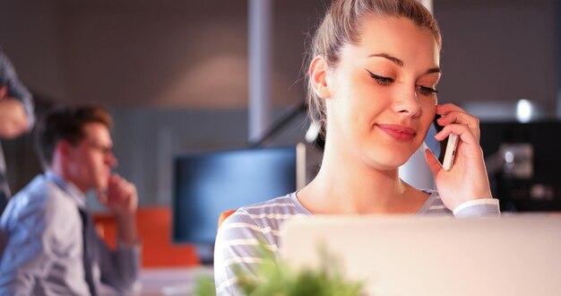 Young woman using mobile phone while working on computer at night in dark office.