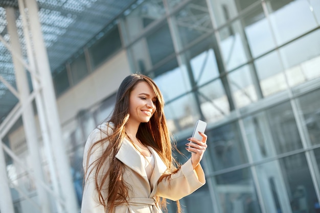 屋外で電話を使用して若い女性。オフィスセンターの近くでスマートフォンとコーヒーのカップを手に持っている美しいビジネス女性の肖像画。