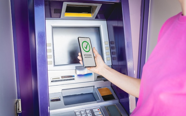 Young woman using the smart phone for cardless withdrawing the cash near the atm