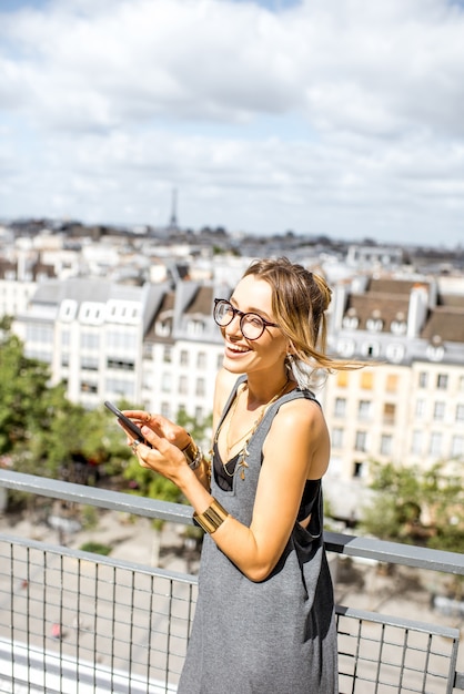 パリの素晴らしい景色を望むテラスに立っているスマートフォンを使用して若い女性