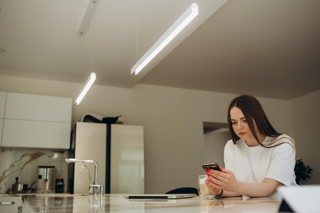 Young woman using smartphone leaning at kitchen table with coffee mug and organizer in a modern home Smiling woman reading phone message Brunette happy girl typing a text message