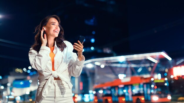 Young woman using smartphone and listening music in earphones while walking in the city at night