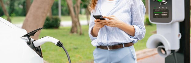 Photo young woman using smartphone online banking application to pay for electric car battery charging from ev charging station during vacation road trip at national park or summer forest panorama exalt