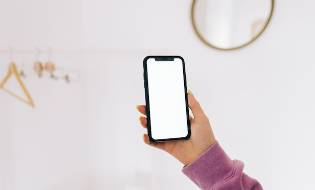 フロアラック付きの白いワードローブの部屋で白い画面のモックアップでスマートフォンを使用している若い女性。
