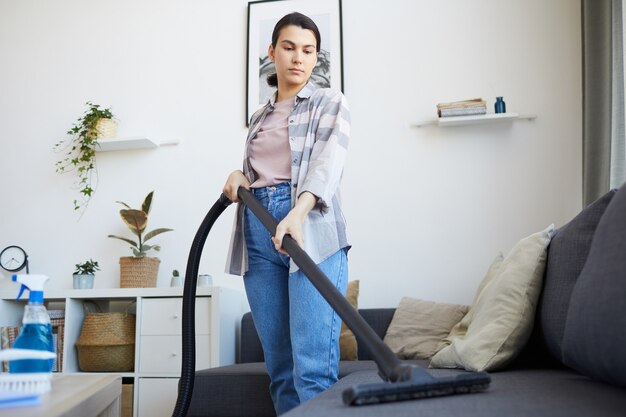 Foto giovane donna che aspira il divano con l'aspirapolvere nel soggiorno