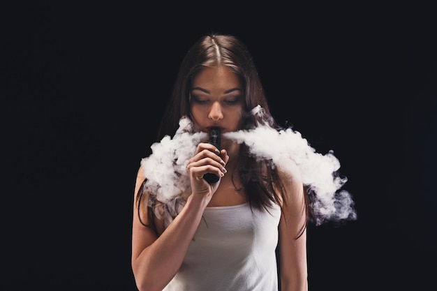 젊은 여자 vaping, 스튜디오 촬영. 검은 배경에 연기 구름을 부는 갈색 머리 소녀. 니코틴 무료 흡연 및 증기 개념, 복사 공간