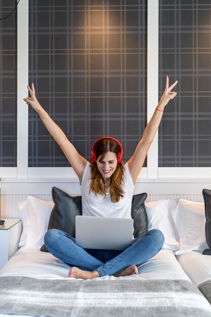 Young woman very happy with her arms up sitting on her bed listening to music and working on her laptop