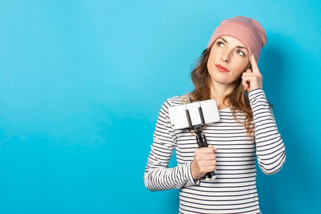 Young woman video blogger holds the phone on a tripod and makes a think gesture on a blue background. The concept of story, vlog, selfie, blog, surprise, shock. Banner.