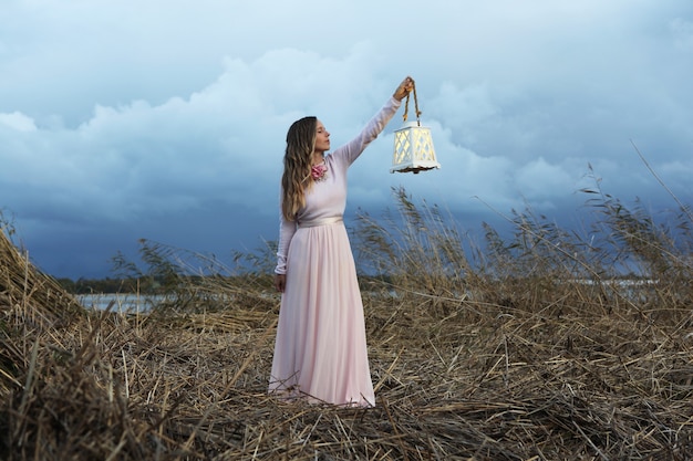 Foto giovane donna in abito lungo rosa vintage in piedi con lanter su erba grigia vicino al mare