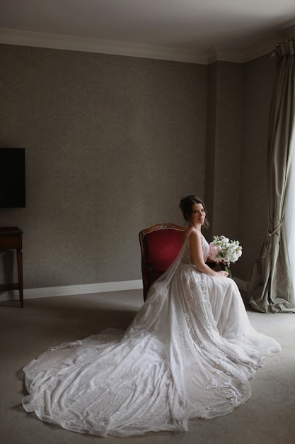 A young woman in a vintage wedding dress with a flower bouquet sits on the chair in the vintage interior