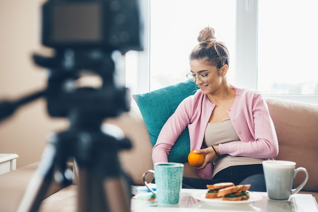 Молодая женщина ведет видеоблог с чашкой чая и бутербродами на столе, носит очки и держит апельсин