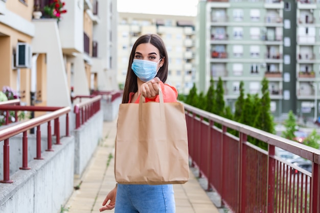 Young woman volunteer with a medical mask is holding a paper bag with food and vegetables. Donation, helping people in quarantine, coronavirus.