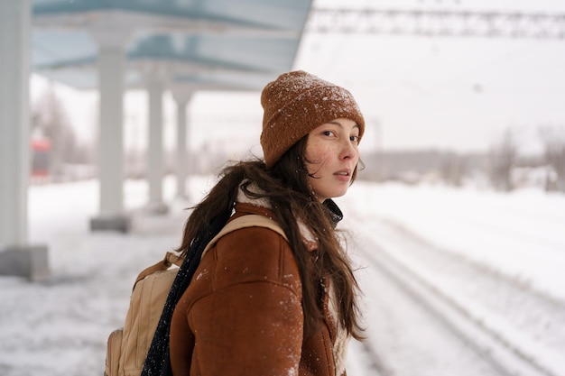 若い女性は、寒い凍るような雪の降る冬の日に屋外の駅に電車が到着するのを待つ