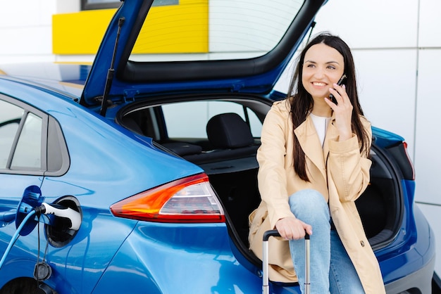 Young woman waiting charging automobile battery from small public station and talking to smartphone while charging automobile