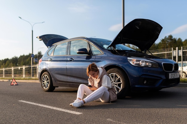 道路上の壊れた車の近くに座って助けを待っている若い女性